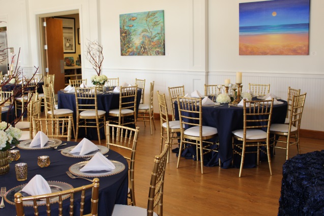 Round tables with navy blue tableclothes, gold chairs, table decorations, and plates. Beach shoreline painting on right side of wall. Fish and aquatic life painting on left side of wall. 